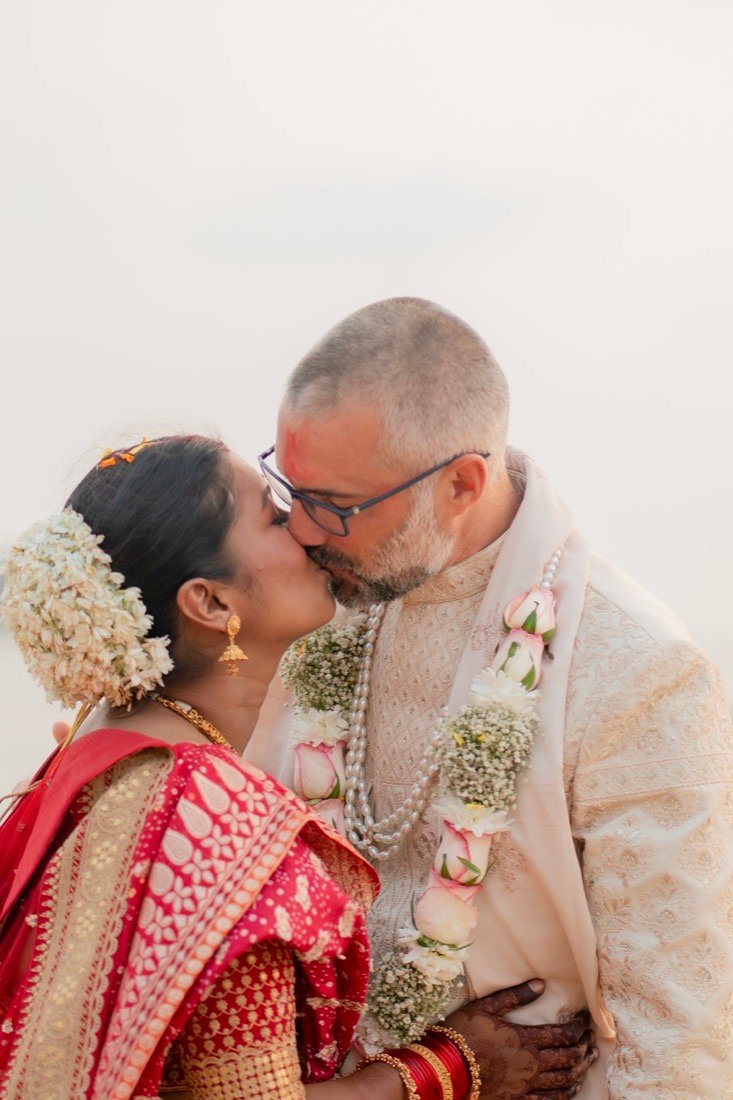 A bride kissing the groom, both garlanded